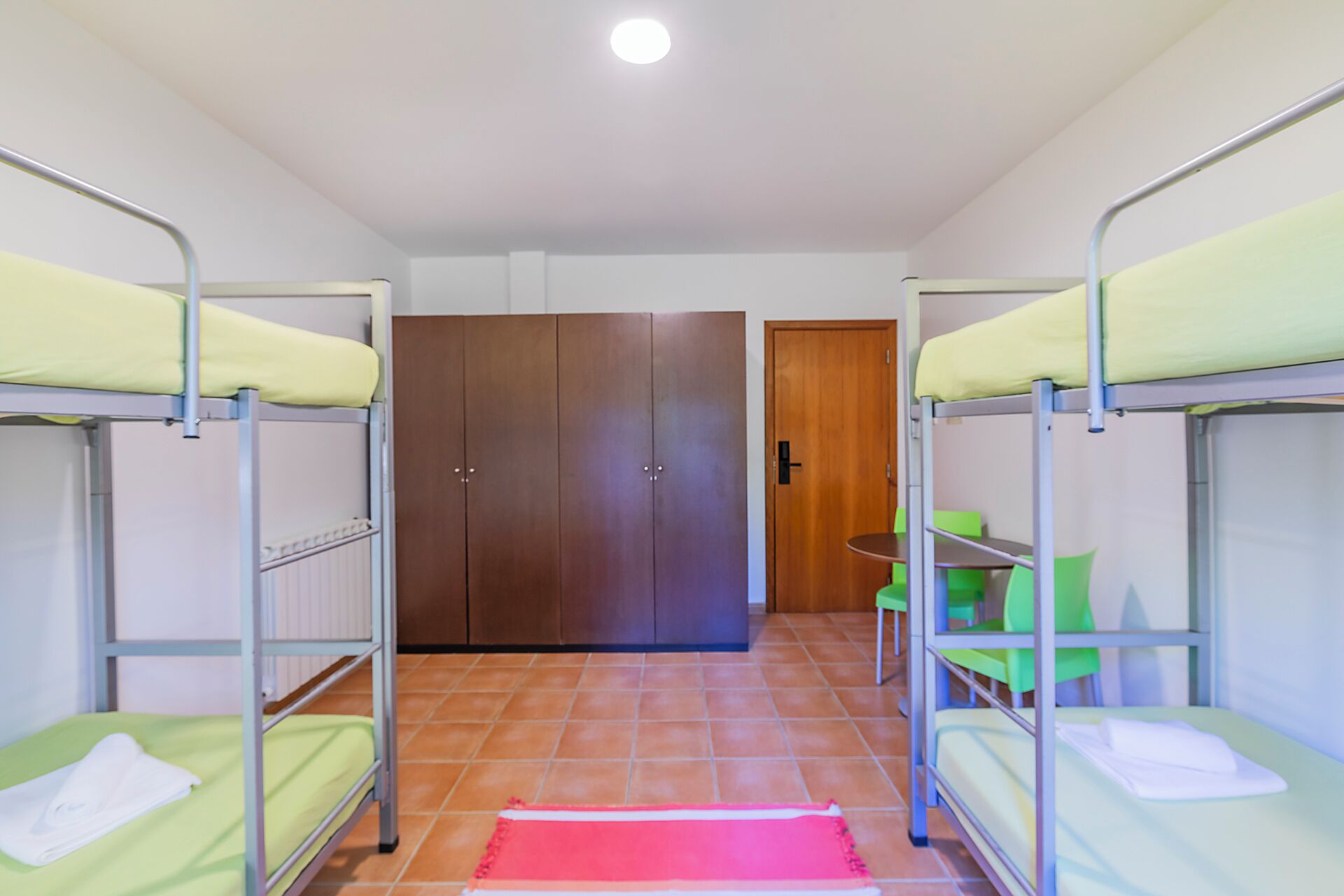 A dormitory room with bunk beds, a wooden wardrobe, a small table with chairs, and a red rug.