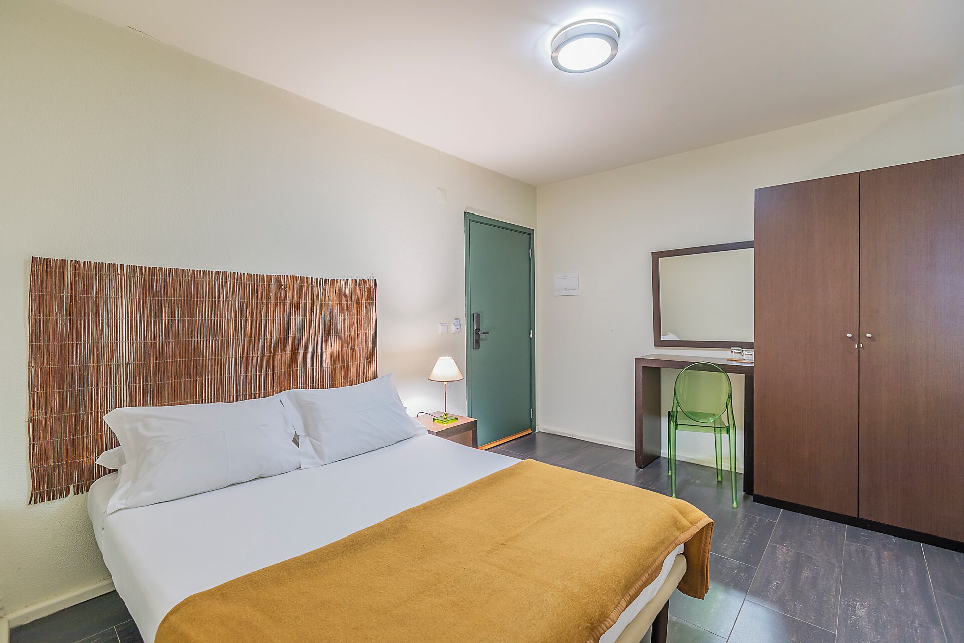 A hotel room with a bed, a desk with a chair, a wardrobe, and a green door.
