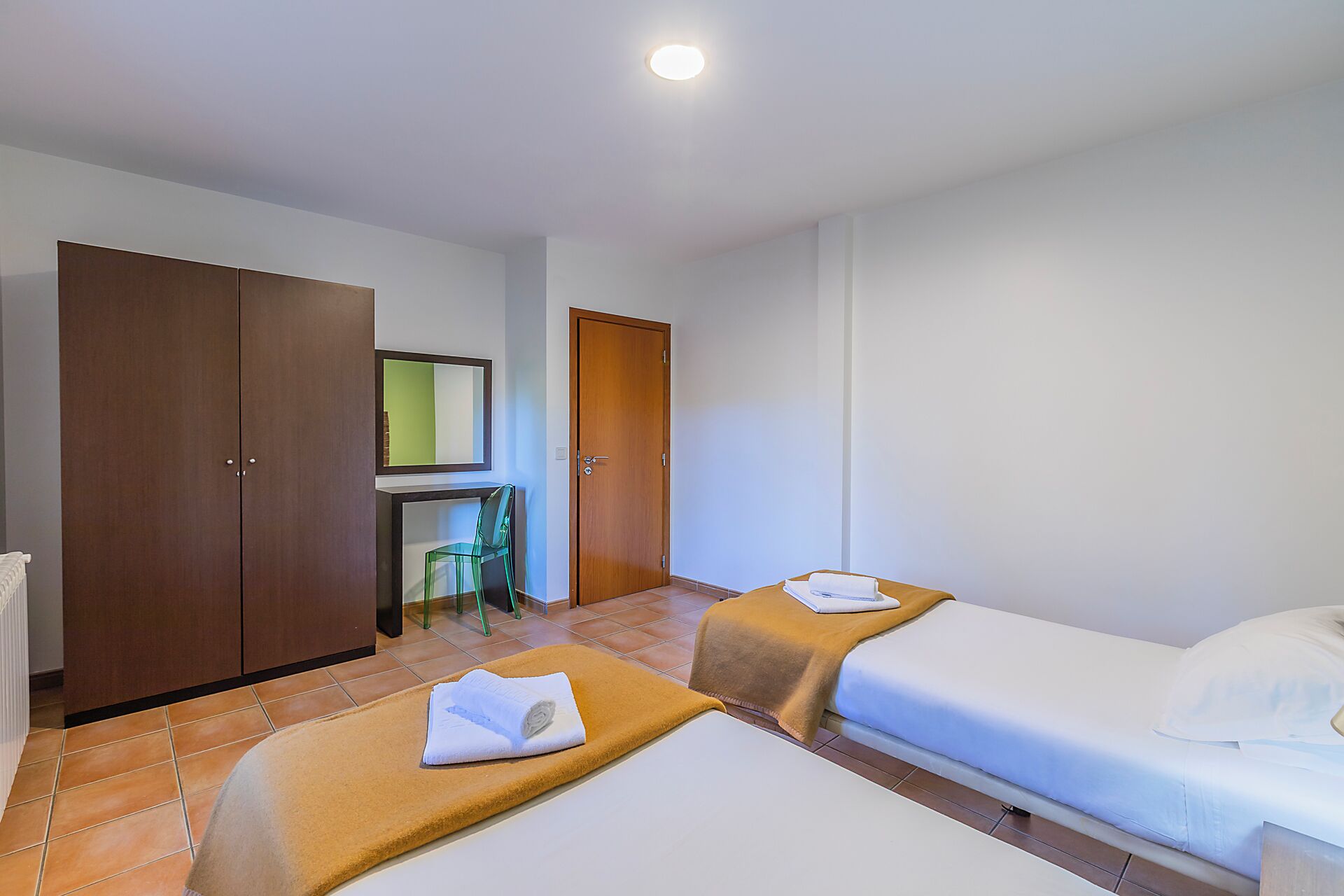 A hotel room with two beds, a wooden wardrobe, a mirror, and a green chair.