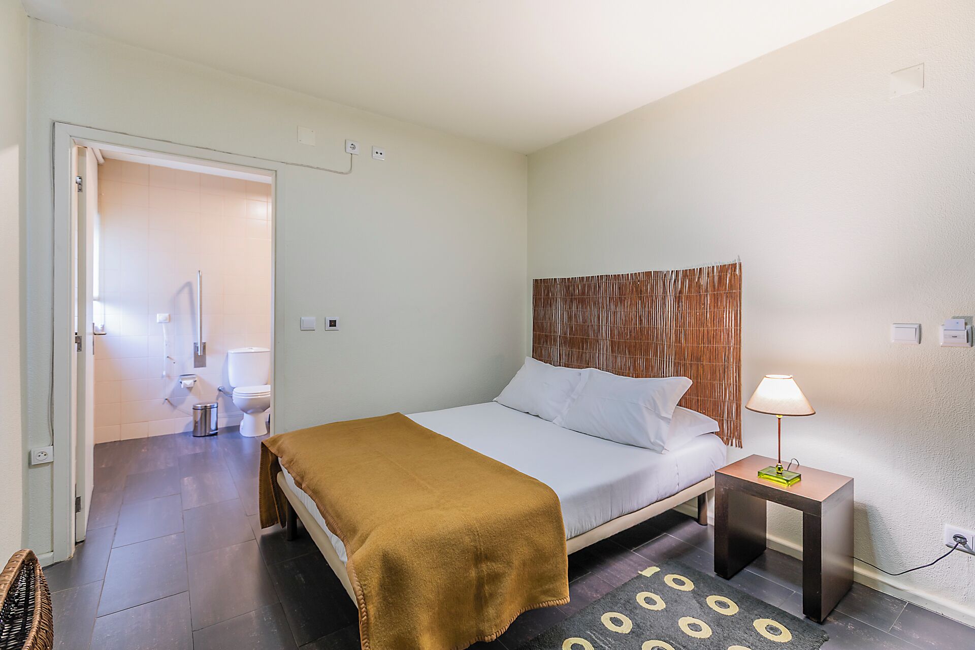 A hotel room with a bed, bedside table, lamp, and a bathroom visible through an open door.