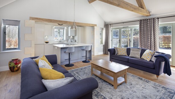 Ein modernes Wohnzimmer mit einem dunkelblauen Sofa, einem Holztisch und einer Küchenzeile mit weißer Arbeitsplatte und Barhocker.