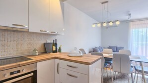 A modern kitchen with a wooden countertop, white cabinets, and a built-in oven. There is a dining area with a table and chairs, and a window with curtains. - Maris apartment (Ližnjan)