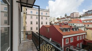 Un balcón con vistas a edificios cercanos, incluyendo un edificio rojo con techo inclinado y un edificio rosa con varias ventanas.