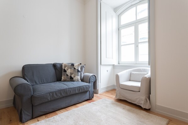 Uma sala de estar moderna com um sofá cinzento, uma poltrona branca e uma grande janela que permite entrada de luz natural.