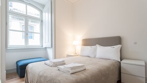 A bedroom with a large bed, a bedside table with a lamp, a bench, and a window with a view of buildings. - FLH Baixa Cozy Apartment II (Lisbon)