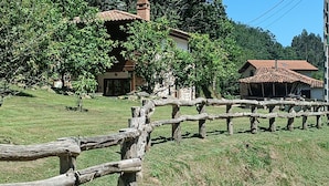 Une clôture en bois rustique, un bâtiment en pierre avec un toit en tuiles et une végétation luxuriante en arrière-plan.