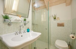 A bathroom with a glass-enclosed shower, a white sink with a blue soap dispenser, and a toilet with a roll of toilet paper. - House Agave (Dubrovnik)