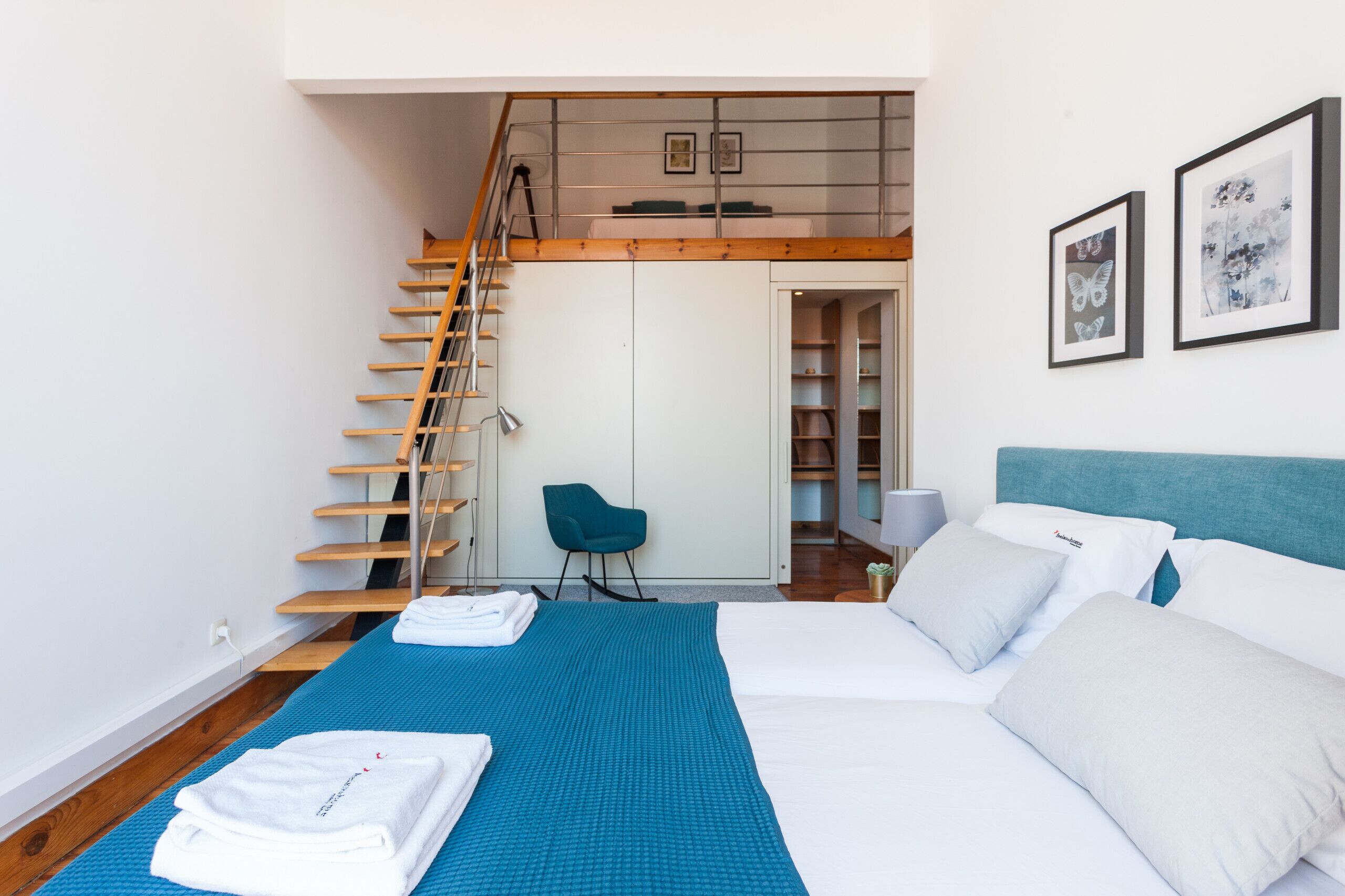 Une chambre d’hôtel moderne avec un grand lit, un tapis bleu, un escalier en bois, une chaise bleue et une armoire.