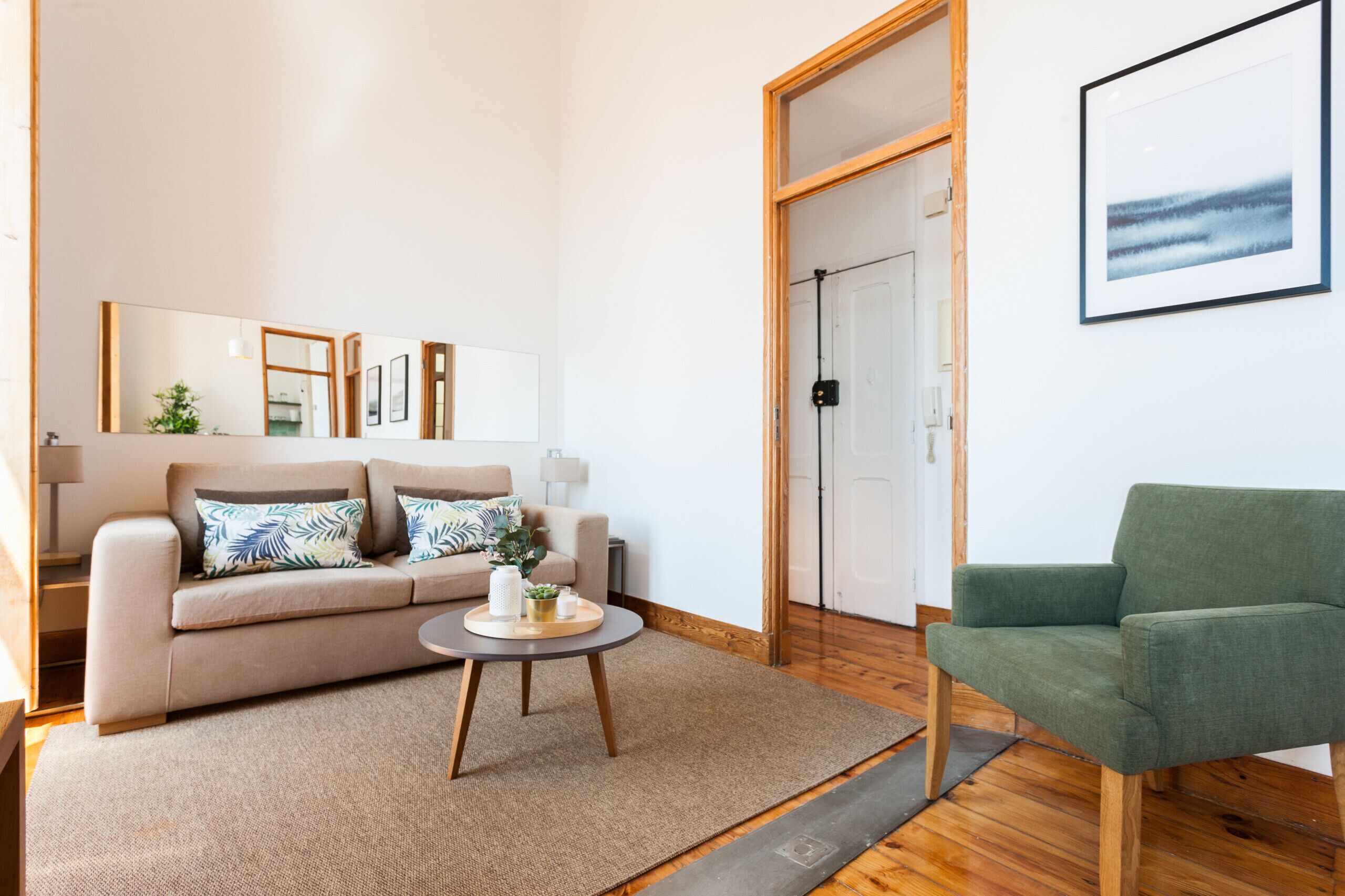 En stue med en beige sofa, en grøn lænestol, et rundt sofabord og et stort spejl på væggen.