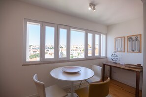 A room with a round table and four chairs, a small wooden desk, and a view of a cityscape through large windows. - FLH Príncipe Real with View (Lisbon)