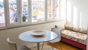 A room with a round table, a sofa, and a view of buildings through large windows. - FLH Príncipe Real with View (Lisbon)