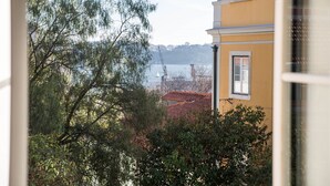 Blick aus einem Fenster auf ein gelbes Gebäude mit rotem Dach, Bäume und einen Kran in der Ferne.