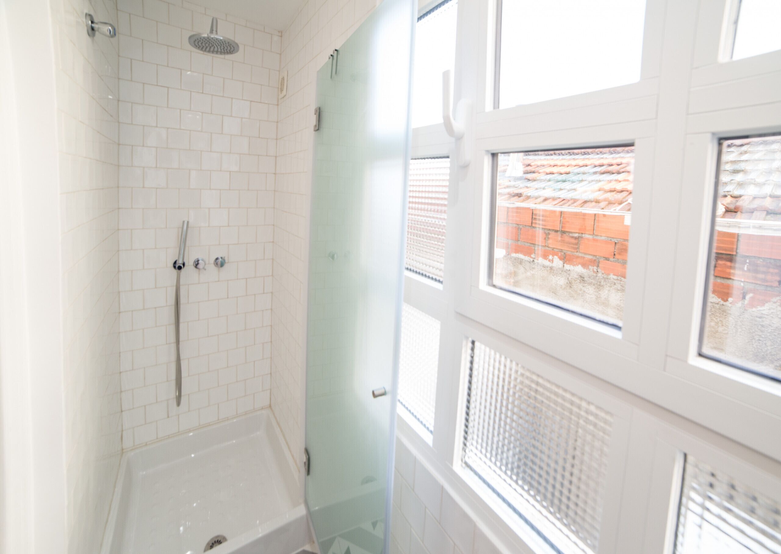 Een badkamer met witte betegelde wanden, een glazen douchecabine, een badkuip en een raam met uitzicht op een bakstenen gebouw.