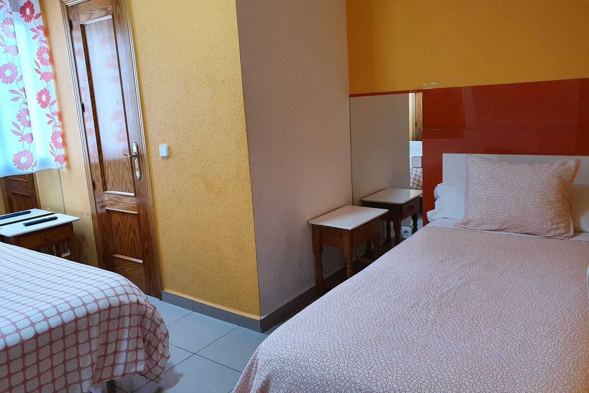 Quarto de hotel com duas camas, uma porta de madeira, uma mesinha e um espelho.