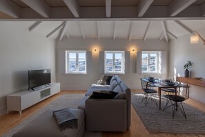 A spacious living area with a large sofa, a dining table with chairs, a TV on a white cabinet, and a view of the city through large windows.