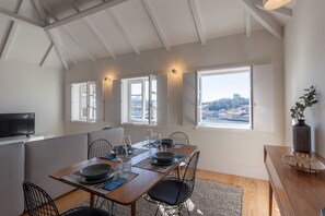 A dining area with a wooden table set for four, a television on the left, and a view of a cityscape through large windows.