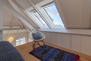 A room with a sloped ceiling, skylights, a white chair with a cushion, a patterned rug, and a wooden floor.