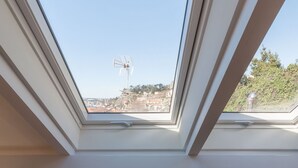 Una habitación con una silla de mimbre blanca, un plaid gris tejido y un tragaluz con vistas a un paisaje urbano.