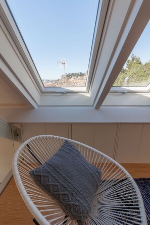 Une pièce avec un fauteuil en osier blanc, un plaid tricoté gris et une lucarne donnant sur un paysage urbain.
