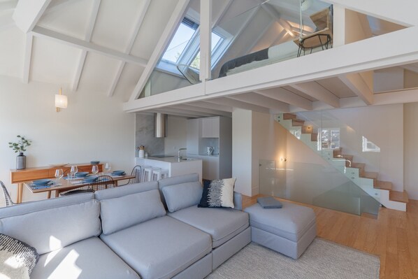 A modern loft apartment with a large sofa, a dining area, a kitchen, and a glass staircase.