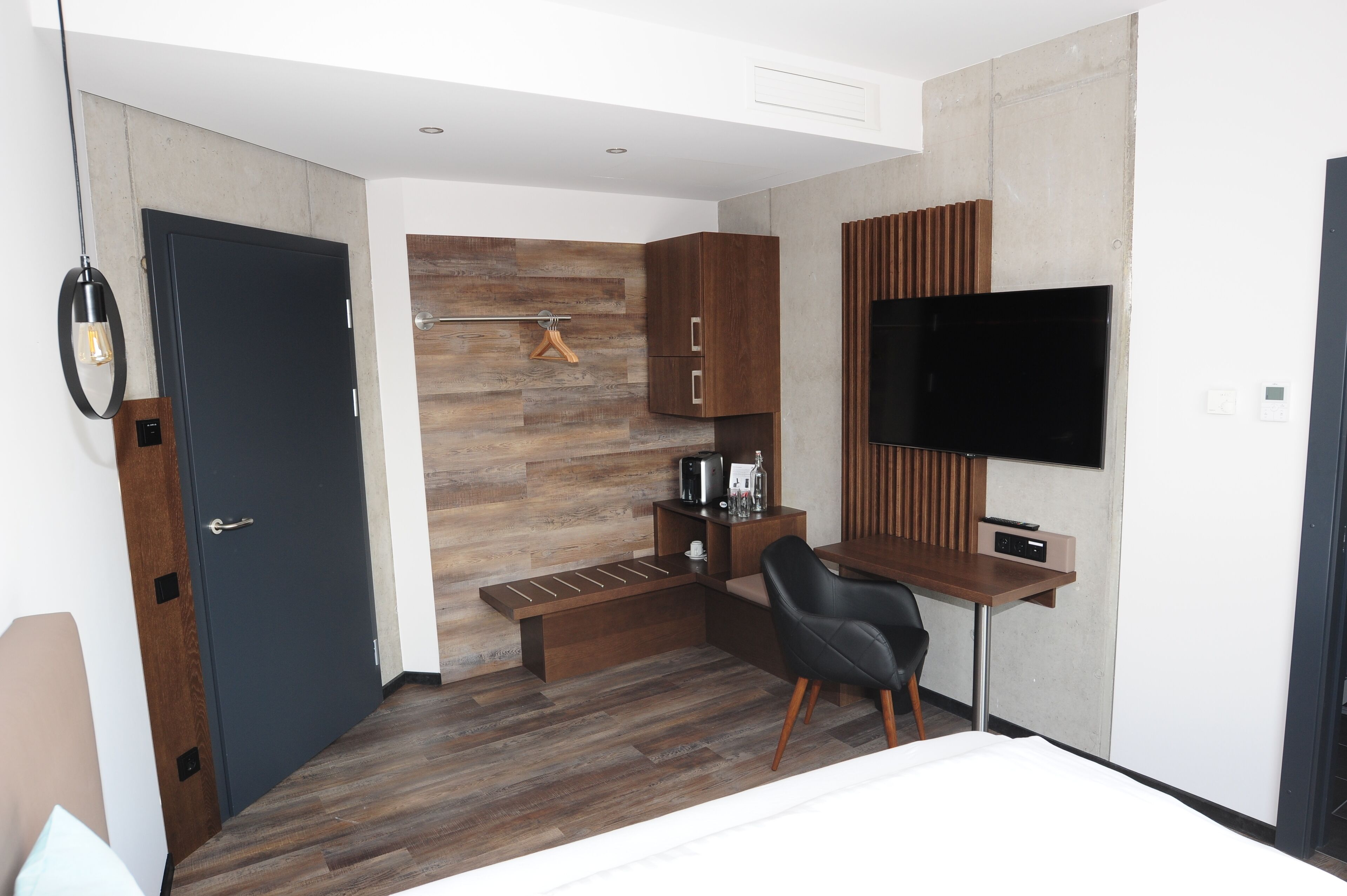 A modern hotel room with a wooden floor, a desk with a chair, a flat-screen TV, and built-in cabinets.