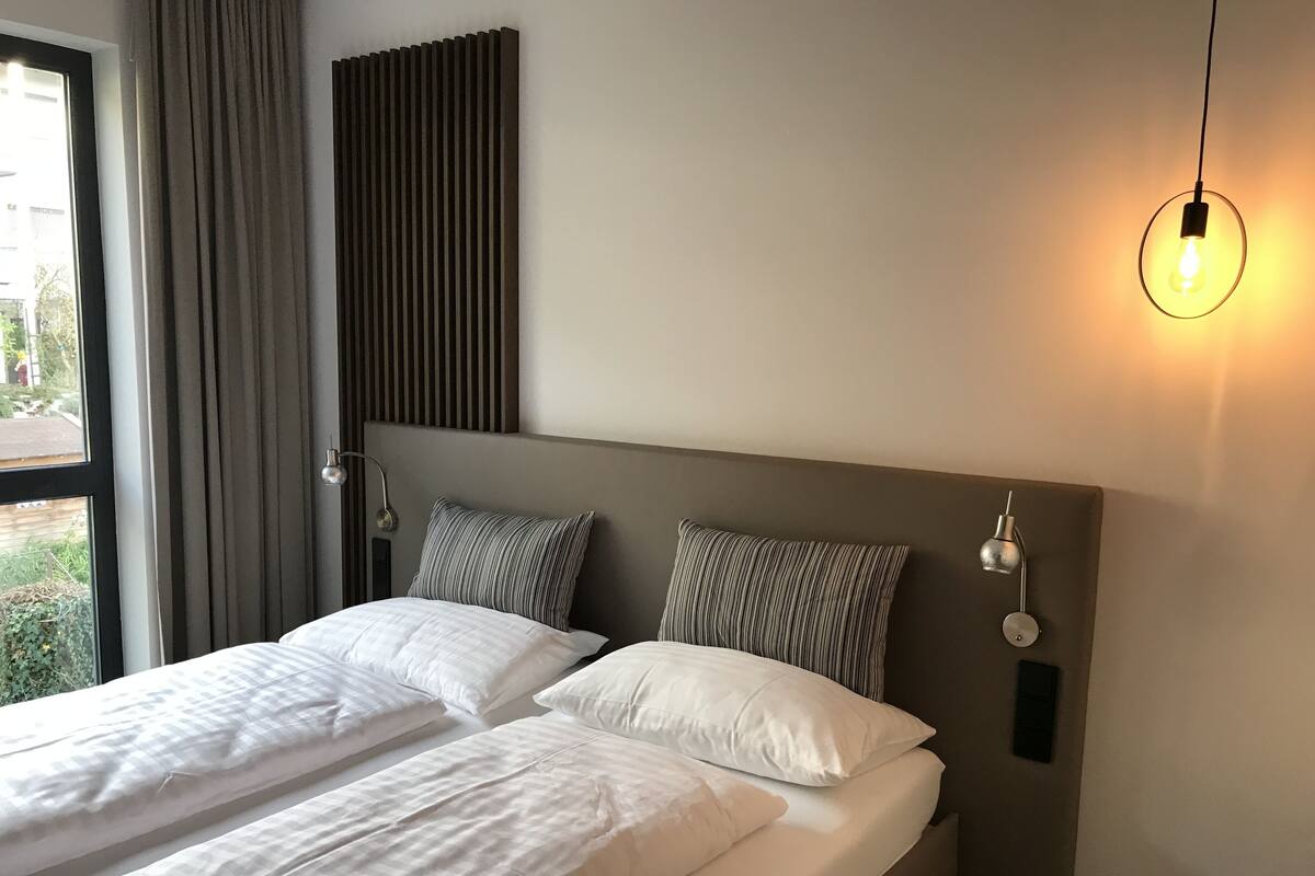 Ein Hotelzimmer mit zwei Betten, einem großen Fenster mit Vorhängen, einer Wandlampe und einer Pendelleuchte.