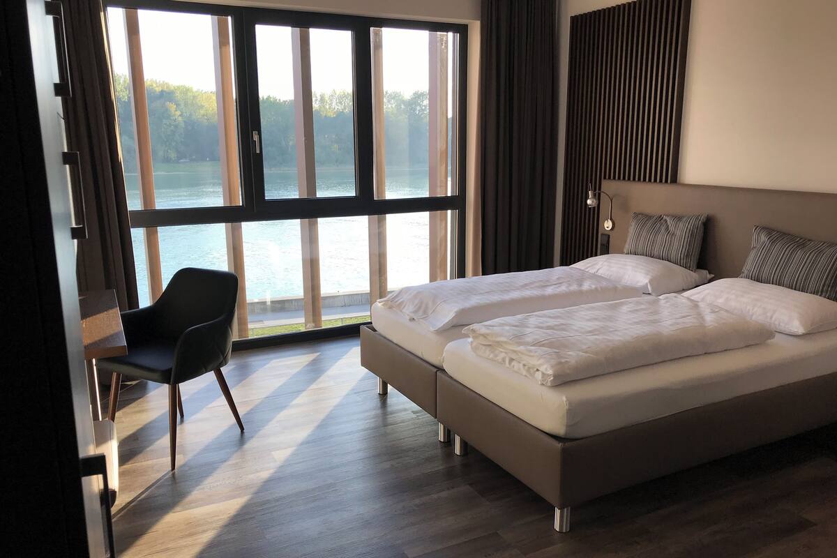 Ein modernes Hotelzimmer mit großem Fenster und Blick auf ein Gewässer, einem Bett mit weißer Bettwäsche, einem Sessel und einer Wandleuchte.