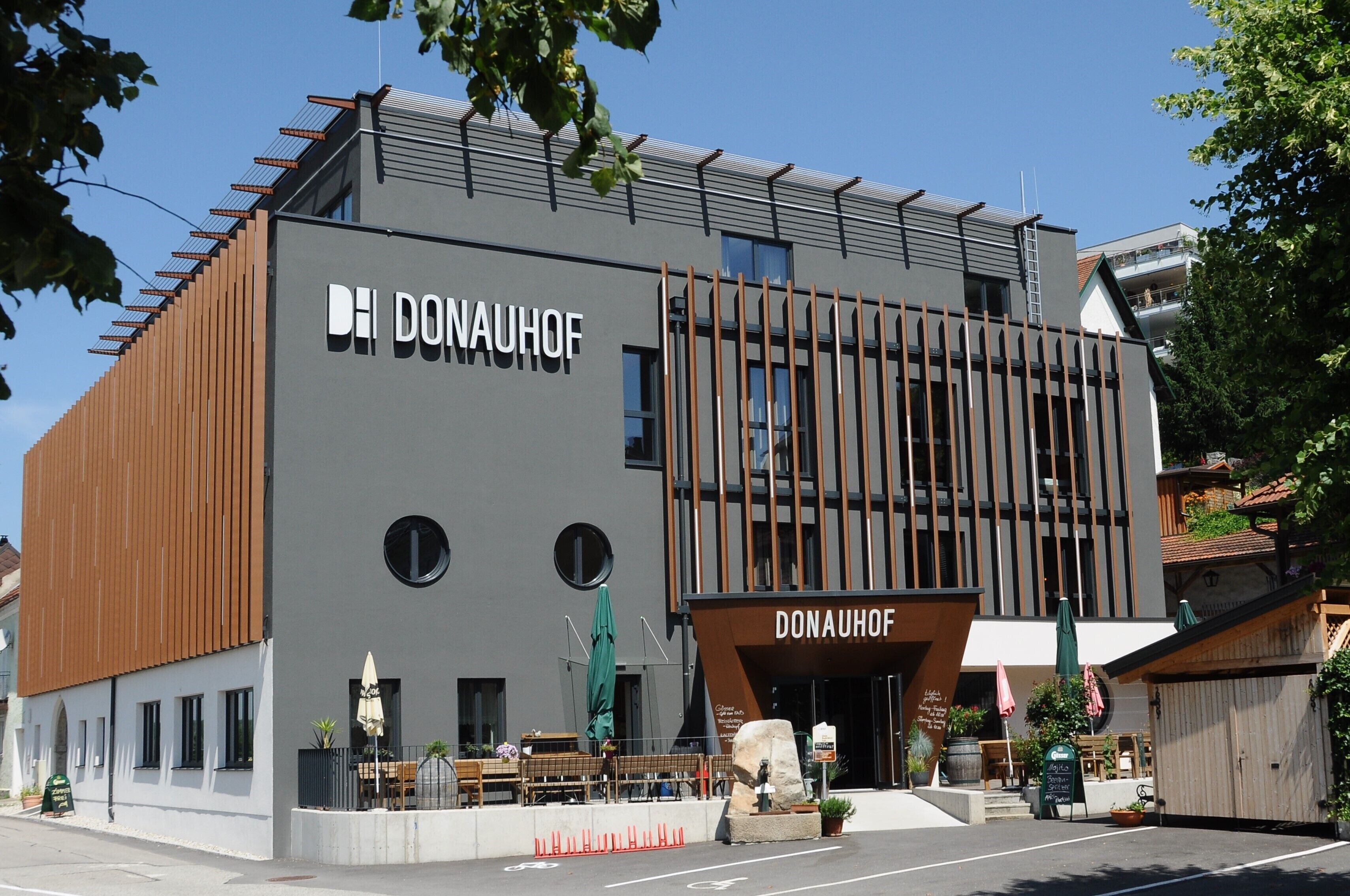 Un edificio moderno con el nombre "DONAUHOF" en la fachada, que combina listones de madera y una pared gris, con mesas y sombrillas en el exterior.
