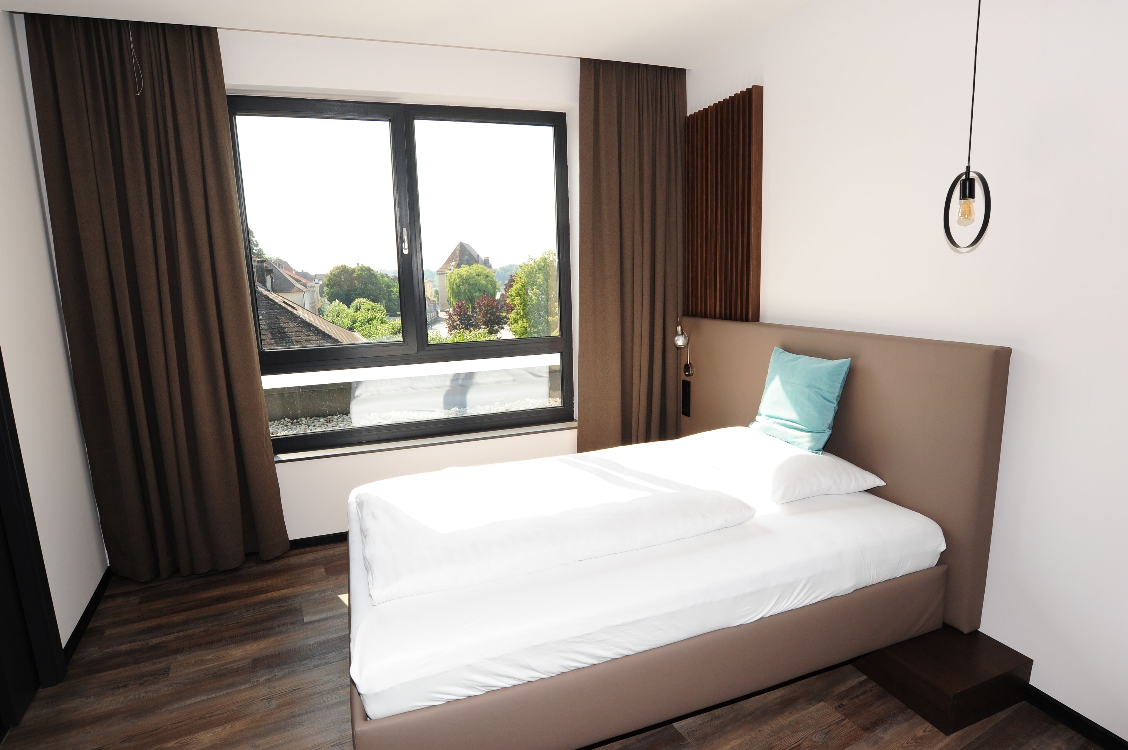 A hotel room with a large window, a bed with white linens, a teal pillow, and a wooden floor.