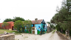 Une route de campagne bordée d’un bâtiment orné d’une fresque colorée, d’un mur de pierres et d’une construction à toit rouge.