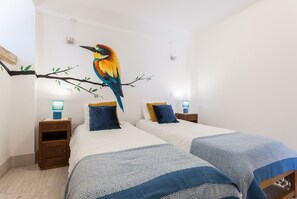A bedroom with two beds, each with a patterned bedspread, a nightstand with a lamp, and a wall mural of a bird on a branch.