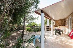 Une terrasse extérieure couverte, dotée d’un mur en pierre, d’un espace barbecue et d’une table avec des chaises.