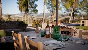 An outdoor dining setup with a table set for four, surrounded by chairs, with a scenic view of hills and trees in the background.
