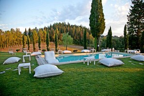 A pool area with lounge chairs and pillows, surrounded by a grassy lawn and trees.