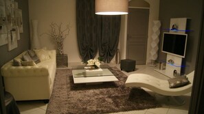 Una sala de estar moderna con un sofá blanco, una mesa de centro de vidrio, un sillón blanco, un televisor y un jarrón decorativo.