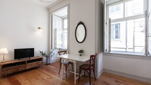 Una stanza con pavimento in legno, un tavolo bianco, due sedie, un televisore su un mobile in legno, uno specchio a parete e una finestra vista edifici.