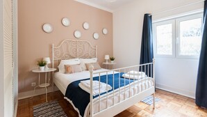 Een slaapkamer met een bed met wit metalen frame, een blauwe spreidek en een wit hoofdbord. Er staan twee nachtkastjes met lampen en een kleine potplant. De kamer heeft een groot raam met donkere gordijnen, en de muren zijn in een zacht perzik kleur geschilderd.