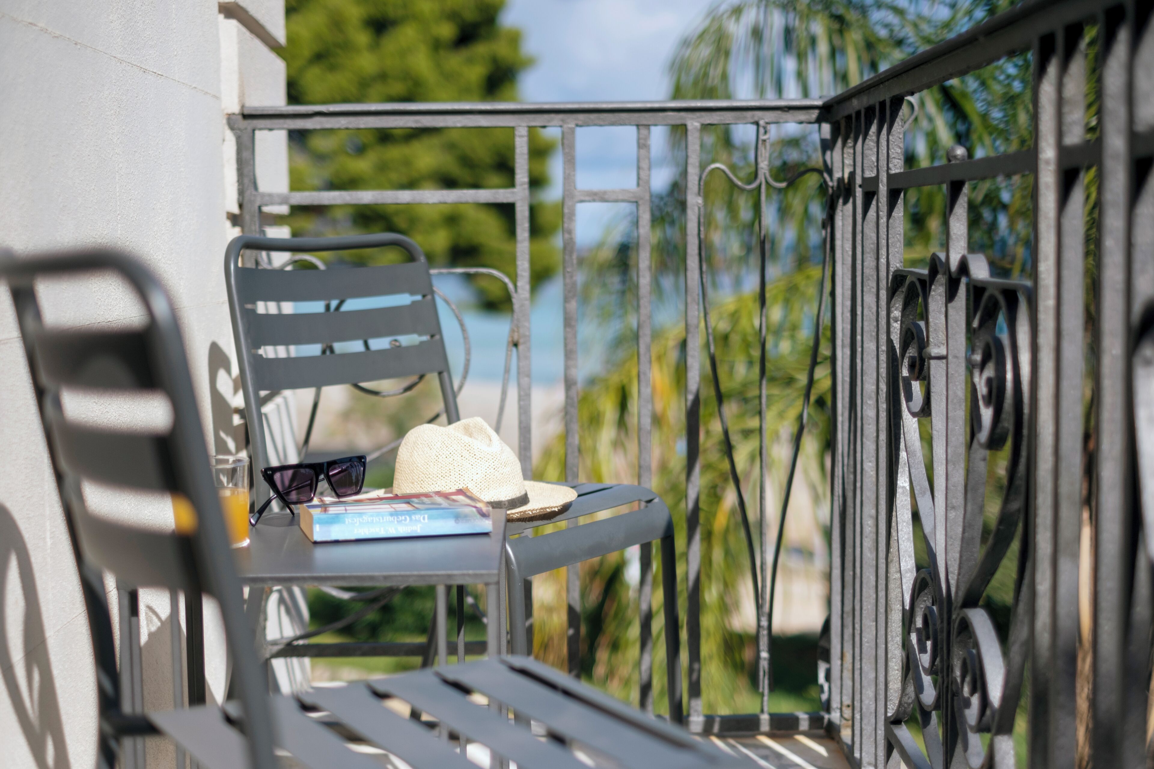 Uma varanda com grades de metal, uma cadeira, um chapéu, óculos de sol e um livro.