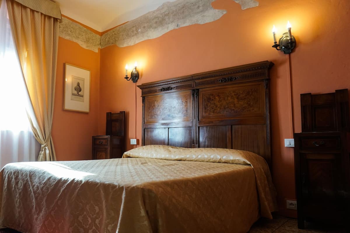 A bedroom with a large bed, wooden headboard, two wall sconces, a framed picture, and a window with curtains.