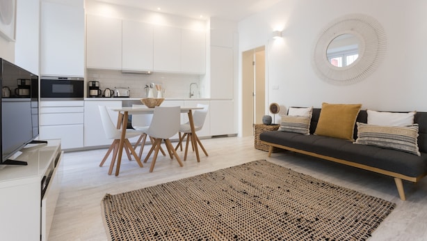 Uma cozinha moderna com armários brancos, uma sala de jantar com mesa e cadeiras, e uma sala de estar com um sofá e um espelho redondo na parede.