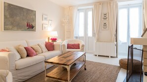 Un salon moderne avec un canapé blanc, une table basse en bois et un fauteuil blanc. Un grand tableau orne le mur, ainsi qu’une lampe et une fenêtre avec des rideaux.