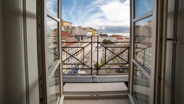 Une vue par une fenêtre ornée d’une grille métallique, donnant sur une rue bordée d’immeubles et sur un ciel dégagé.