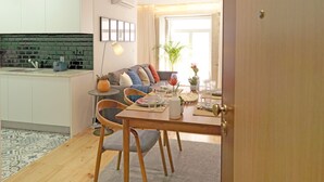 A modern kitchen with a built-in oven and sink, a dining area with a wooden table and chairs, and a living room with a sofa and a plant.