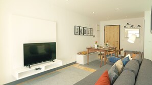 A modern living room with a flat-screen TV on a white stand, a dining table with chairs, a sofa with cushions, and a wooden door.