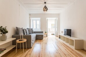 Un salon moderne avec un canapé d’angle gris, une petite chaise en bois, un téléviseur posé sur une étagère blanche et une plante en pot.