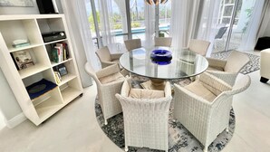 Een moderne woonkamer met een glazen tafel, omringd door rieten stoelen, een boekenkast en uitzicht op een zwembad.