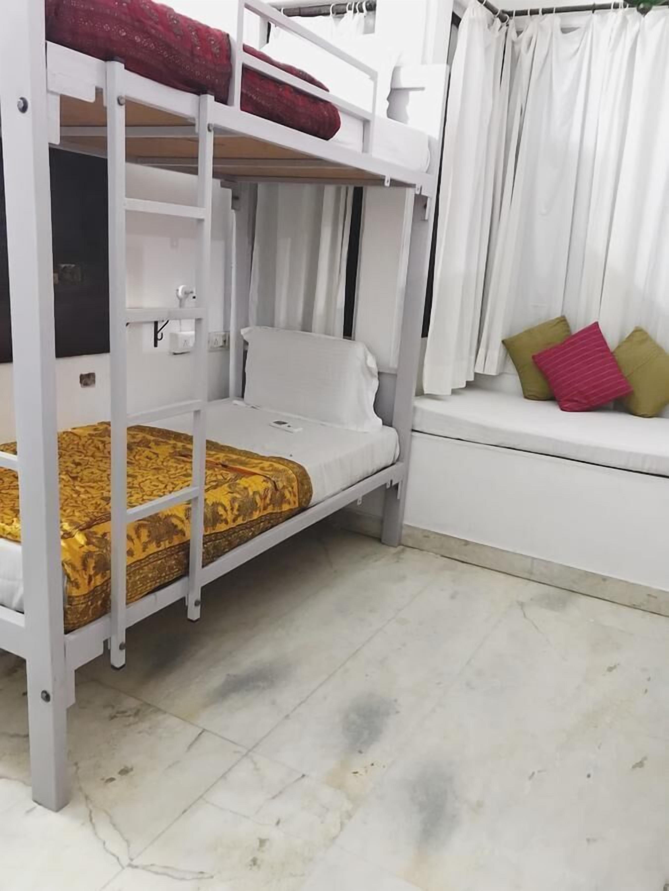 A bunk bed with a lower bunk covered in a patterned blanket, a white mattress on the upper bunk, and a window with white curtains.