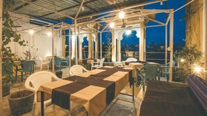 Un área de comedor al aire libre con mesas y sillas, ventanas amplias y vistas de la ciudad por la noche.