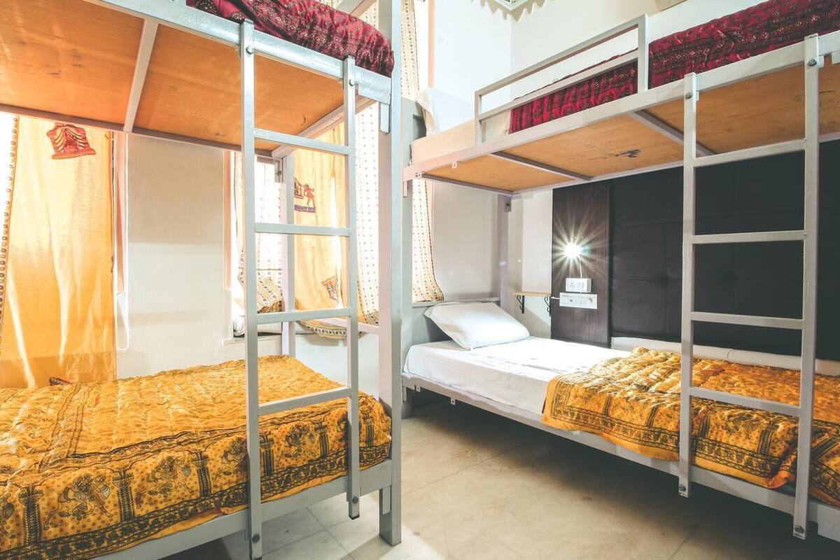 A dormitory room with bunk beds, a bedside table with a lamp, and curtains on the windows.