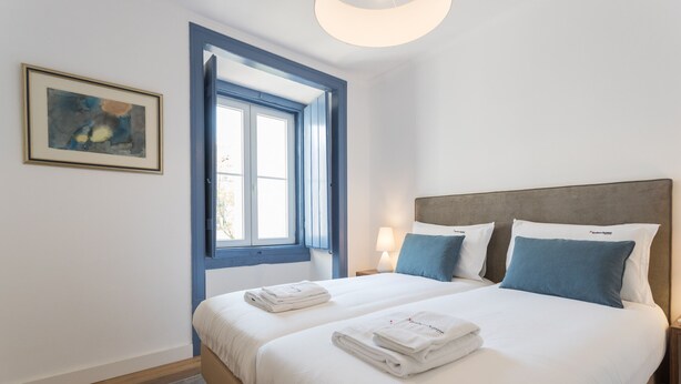 A double bed with white linens and blue pillows, a framed abstract painting on the wall, and a window with blue trim.